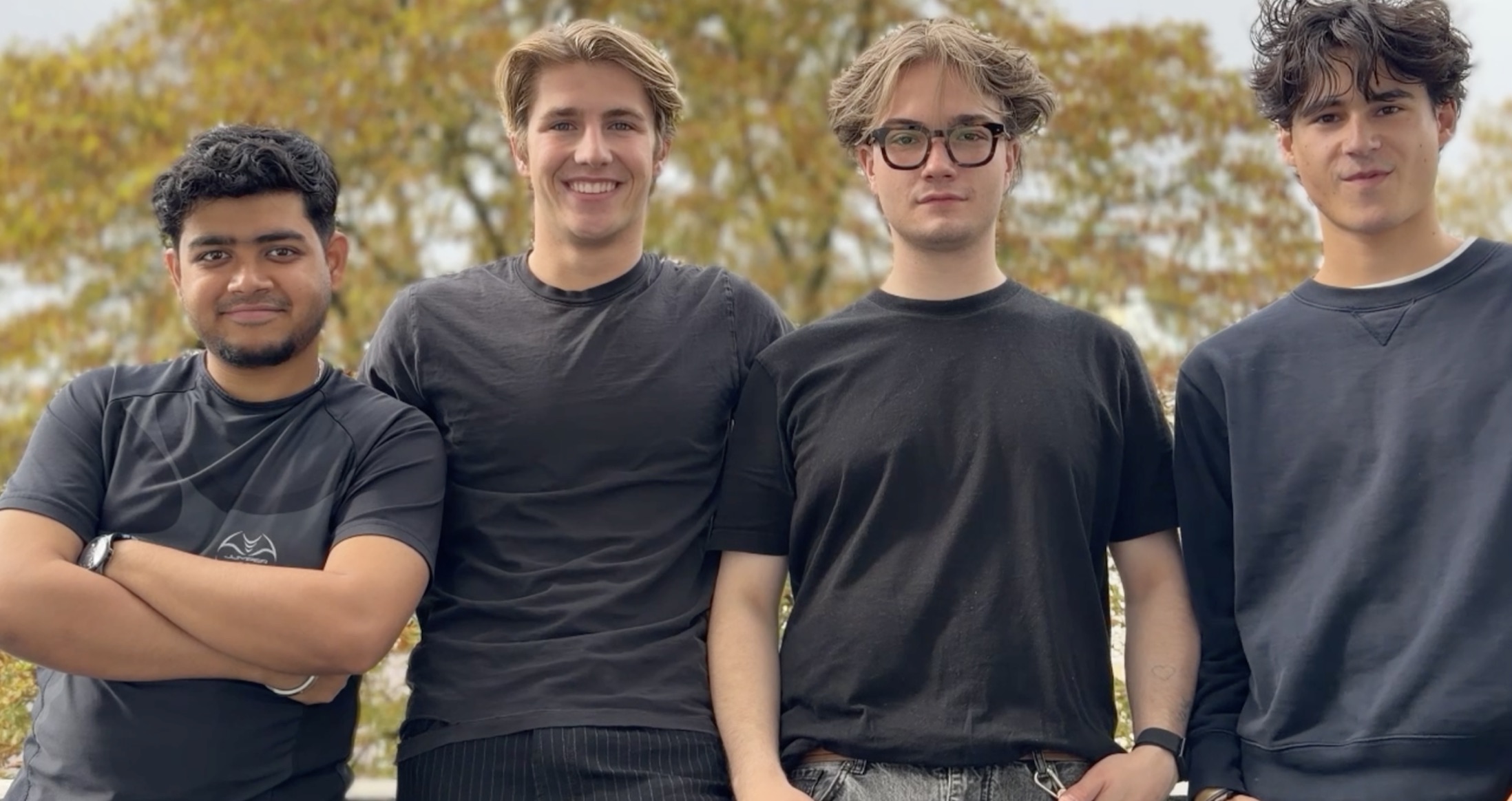 Das Team von SEE:ME: vier Personen in dunkler Kleidung stehen nebeneinander im Freien, im Hintergrund ein Baum mit Herbstlaub.